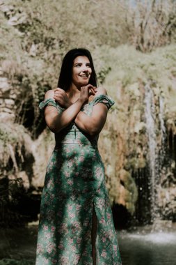 Bir dağ şelalesinin yanında poz veren turkuaz elbiseli güzel bir manken. Vahşi yaşam kavramı.