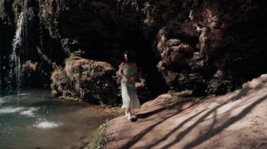 Bir dağ şelalesinin yanında poz veren turkuaz elbiseli güzel bir manken. Vahşi yaşam kavramı.