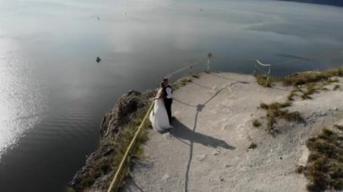 Deniz kıyısındaki bir uçurumun kenarında düğün. Gelin ve damat yüksek uçurumun kenarında duruyorlar. Günbatımı ışığının 4k hava aracı videosu