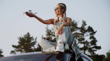 Gözlüklü şık bir kadın gün batımında bir arabanın çatısında oturan bir drone fırlatıyor. Drone pilotu konsepti