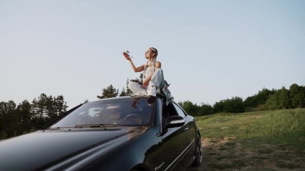 戴眼镜的时髦女人在日落时坐在车顶上发射无人驾驶飞机 无人机试点概念 — 图库视频影像