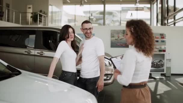 自動車販売センターで 若い家族に新しい自動車を販売する車の販売員 車のショールームで美しい若いカップルは購入する新しい車を選択します — ストック動画