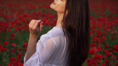 Haşhaş tarlasında kısa beyaz bir elbise giymiş güzel bir kadın, güzel kırmızı çiçeklerin arasında, gülümseyen ve rüzgarda poz veren. Doğa konsepti.