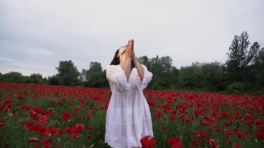 Haşhaş tarlasında kısa beyaz bir elbise giymiş güzel bir kadın, güzel kırmızı çiçeklerin arasında, gülümseyen ve rüzgarda poz veren. Doğa konsepti.