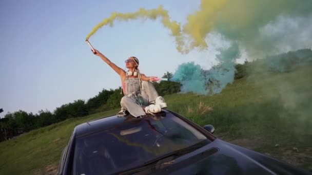 煙爆弾を持つ若いスタイリッシュな女性は車の屋根の上に座って 日没を楽しんでいます — ストック動画