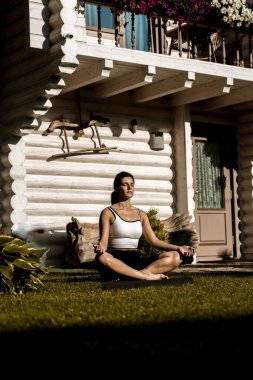 Formda, spor elbiseli güzel bir kadın evinin yanındaki yeşil çimlerin üzerinde lotus pozisyonunda otururken meditasyon yapıyor. Evde yoga egzersizi yapan genç bir kadın. 