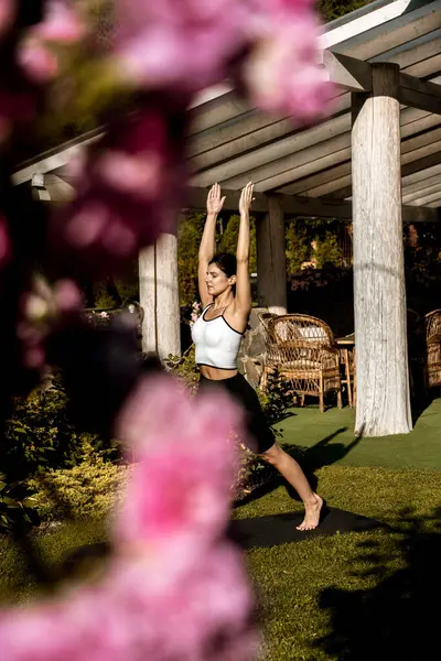 Formda, spor elbiseli güzel bir kadın evinin yanındaki yeşil çimlerin üzerinde lotus pozisyonunda otururken meditasyon yapıyor. Evde yoga egzersizi yapan genç bir kadın. 