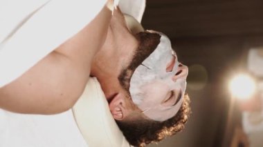 Masaj odasında masaj ve yüz bakımı yaptıran genç bir adam. Güzellik salonunda yüz masajı yaptıran yakışıklı bir adam. Yüz masajı ve cilt bakımı konsepti.