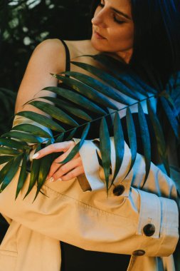 Tropik bir bahçede poz veren bej renkli trençkotlu seksi kadın. Moda çekim konsepti