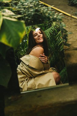 Tropik bir bahçede poz veren bej renkli trençkotlu seksi kadın. Moda çekim konsepti