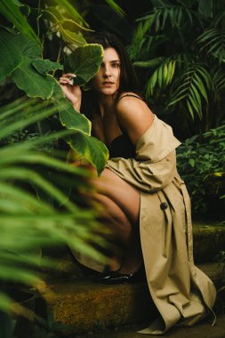 Tropik bir bahçede poz veren bej renkli trençkotlu seksi kadın. Moda çekim konsepti