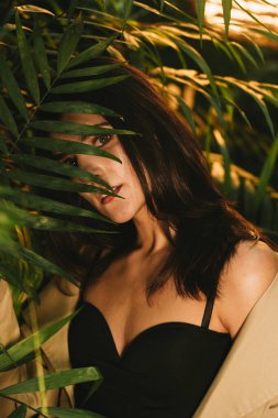 Tropik bir bahçede poz veren bej renkli trençkotlu seksi kadın. Moda çekim konsepti