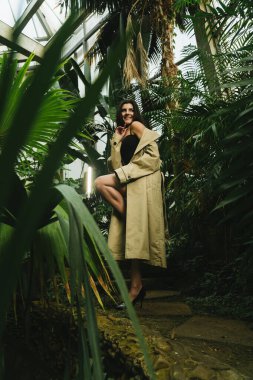 Tropik bir bahçede poz veren bej renkli trençkotlu seksi kadın. Moda çekim konsepti