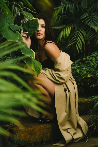 Sexy woman in a beige trench coat posing in a tropical garden. Fashion shooting concept