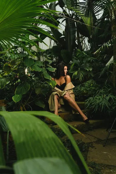 Sexy woman in a beige trench coat posing in a tropical garden. Fashion shooting concept