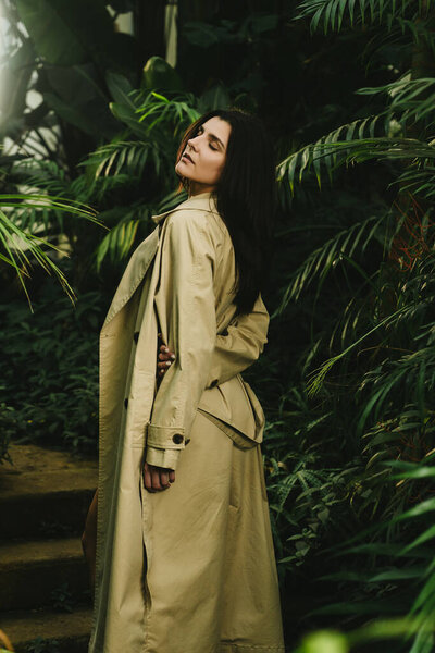 Sexy woman in a beige trench coat posing in a tropical garden. Fashion shooting concept