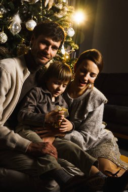 Mutlu aile babası, anne ve oğlu yeni yıl ağacını süslüyor, eğleniyor, dans ediyor ve hediyeleri açıyor. Noel ve Yeni Yıl Arifesi kavramı. Dikey çekim