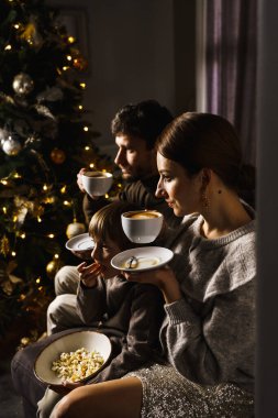 Mutlu aile Noel 'i kutlar, film izler ve yılbaşı ağacının yanında patlamış mısır ve pizza yer. Dikey fotoğraf