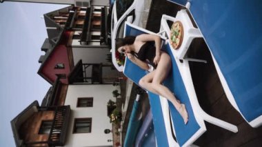 Siyah mayo giyen seksi kadın havuz kenarında suşi ve pizza yiyor. Otel tatili konsepti. Dikey video