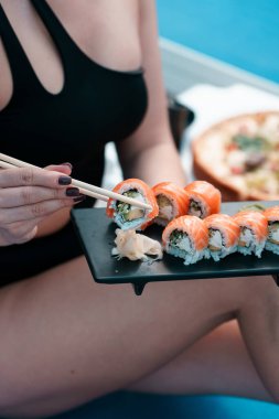 A young woman in a black swimsuit eats a Philadelphia roll near the pool. Concept of rolls and sushi. Relaxation near the pool. Summer season