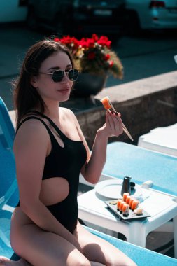 A young woman in a black swimsuit eats a Philadelphia roll near the pool. Concept of rolls and sushi. Relaxation near the pool. Summer season