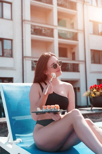 A young woman in a black swimsuit eats a Philadelphia roll near the pool. Concept of rolls and sushi. Relaxation near the pool. Summer season