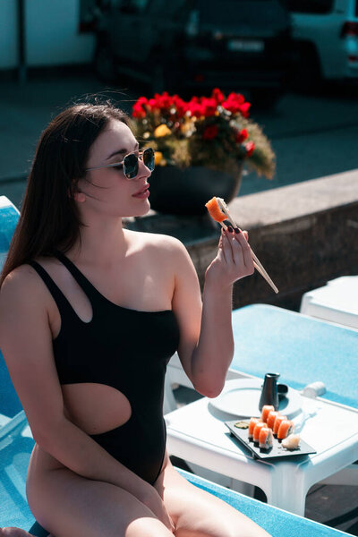 A young woman in a black swimsuit eats a Philadelphia roll near the pool. Concept of rolls and sushi. Relaxation near the pool. Summer season