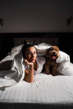 Genç bir kadın yatakta oyuncak köpekle oynuyor ve poz veriyor. Hayvanlarla arkadaşlık kavramı.