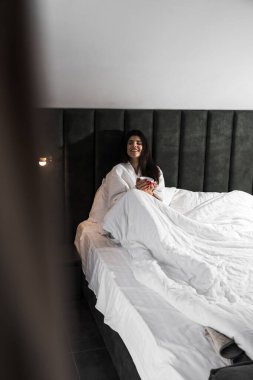 Hoş bir kadın bir otel odasında uyanıyor ve kışın pencereden manzaranın tadını çıkarıyor. Yatakta kahve. Otel tatil konsepti