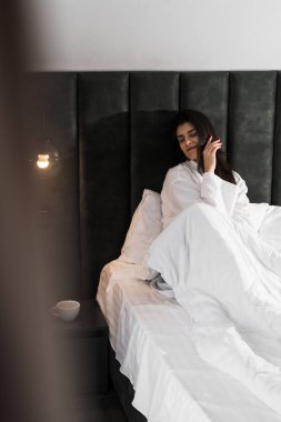 Hoş bir kadın bir otel odasında uyanıyor ve kışın pencereden manzaranın tadını çıkarıyor. Yatakta kahve. Otel tatil konsepti