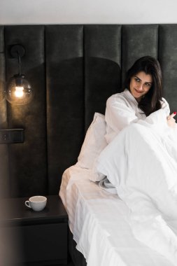 Hoş bir kadın bir otel odasında uyanıyor ve kışın pencereden manzaranın tadını çıkarıyor. Yatakta kahve. Otel tatil konsepti