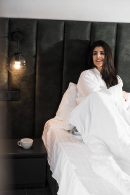 Hoş bir kadın bir otel odasında uyanıyor ve kışın pencereden manzaranın tadını çıkarıyor. Yatakta kahve. Otel tatil konsepti