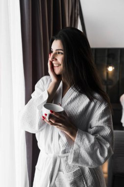 Hoş bir kadın bir otel odasında uyanıyor ve kışın pencereden manzaranın tadını çıkarıyor. Yatakta kahve. Otel tatil konsepti