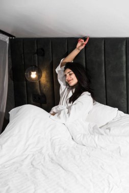 Hoş bir kadın bir otel odasında uyanıyor ve kışın pencereden manzaranın tadını çıkarıyor. Yatakta kahve. Otel tatil konsepti