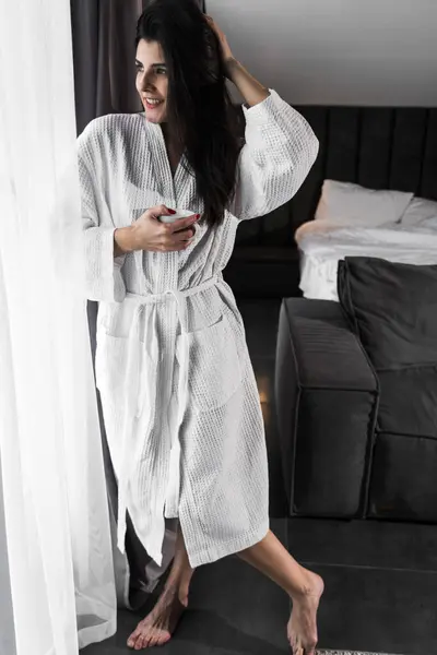 Hoş bir kadın bir otel odasında uyanıyor ve kışın pencereden manzaranın tadını çıkarıyor. Yatakta kahve. Otel tatil konsepti