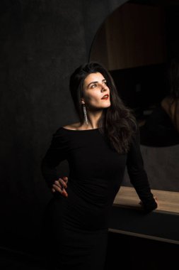 Siyah elbiseli seksi kadın karanlık bir odada aynanın yanında poz veriyor. Moda çekim konsepti