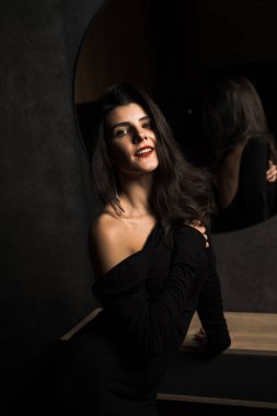 Siyah elbiseli seksi kadın karanlık bir odada aynanın yanında poz veriyor. Moda çekim konsepti
