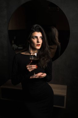 Siyah elbiseli lüks kadın aynanın yanında bir kadeh şarapla poz veriyor.