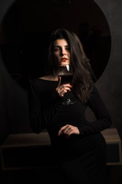 Siyah elbiseli lüks kadın aynanın yanında bir kadeh şarapla poz veriyor.
