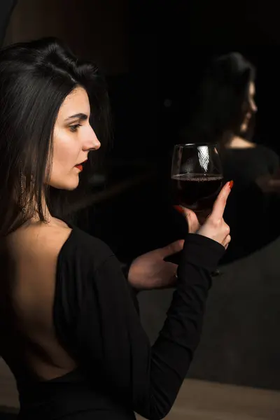 Luxurious woman in a black dress posing with a glass of wine near the mirror