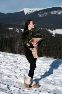 Güneş gözlüklü güzel bir kadın kozalaklı bir buketle kar örtülü dağların arkasında poz veriyor.