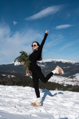 Güneş gözlüklü güzel bir kadın kozalaklı bir buketle kar örtülü dağların arkasında poz veriyor.