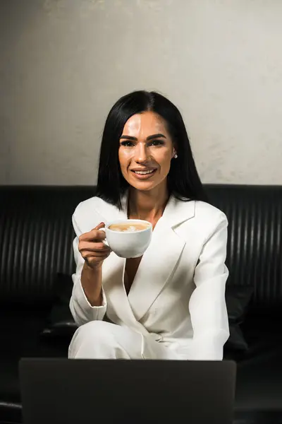 A business woman in a white suit works at a laptop in the office and drinks coffee. Office work concept. Remote work on a PC