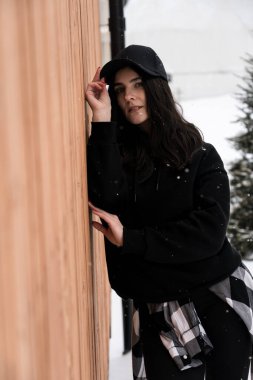 Siyah şapkalı genç bir kadın bir kulübenin yakınındaki karlı dağların arka planında poz veriyor. Kış tatili kavramı