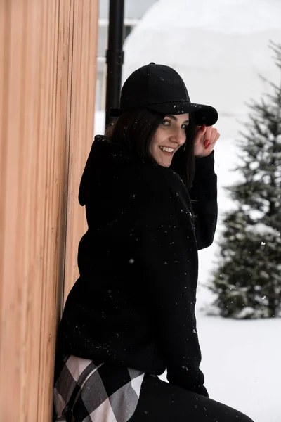 A young woman in a black cap poses against the backdrop of snow-capped mountains near a cottage. Winter holiday concept