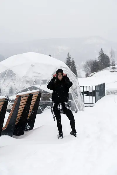 A young woman in a black cap poses against the backdrop of snow-capped mountains near a cottage. Winter holiday concept