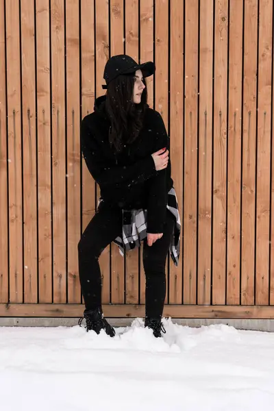 A young woman in a black cap poses against the backdrop of snow-capped mountains near a cottage. Winter holiday concept