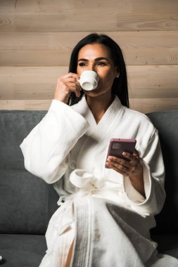 Beyaz bornozlu genç bir kadın ahşap bir otel odasında espresso içiyor. Otel tatili konsepti. Sabah kahvesi