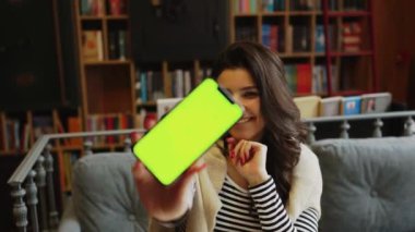 Genç bir kadın kahve dükkanında kahve içiyor ve telefonda çalışıyor. Kadın, telefon ekranını sarıyor. Telefonda yeşil ekran. Serbest konsept 4K