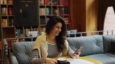 Genç bir kadın kahve dükkanında kahve içiyor ve telefonda çalışıyor. Kadın, telefon ekranını sarıyor. Telefonda yeşil ekran. Serbest konsept 4K